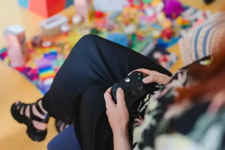 Eine Frau hält einen Gamecontroller in den Händen und spielt vor einem leicht verschwommenen, bunt beleuchteten Hintergrund.
