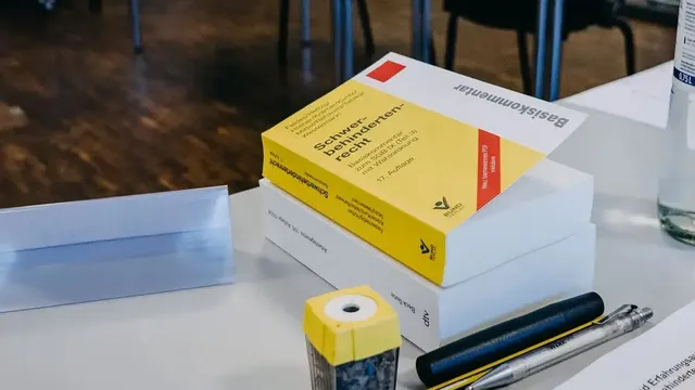 Auf einem Tisch liegen ein dicker gelber Gesetzeskommentar, ein weißes Buch, ein Bleistift, zwei Stifte und ein gelber Spitzer. Im Hintergrund stehen Tische und Stühle eines Seminarraums.