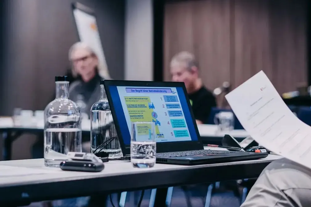 Laptop auf einem Konferenztisch zeigt eine Präsentationsfolie mit dem Titel „Der Begriff der Mitbestimmung", daneben Wasserflaschen und ein Glas.