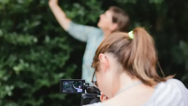 Person filmt mit einer Videokamera eine andere Person vor grünem Buschwerk, Kameradisplay zeigt die Aufnahme.