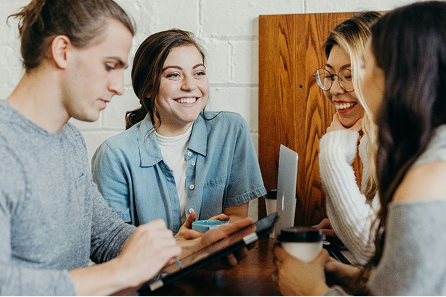 Vier Personen sitzen an einem Tisch zusammen. Auf dem Tisch stehen ein Laptop, ein Tablet und eine Kaffeetasse.