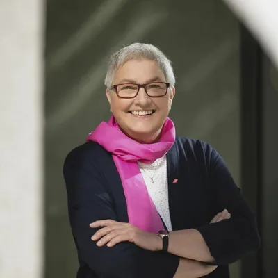 Lächelnde Frau (Elke Hannack) mit Brille und pinkem Schal vor neutralem Hintergrund, mit verschränkten Armen.