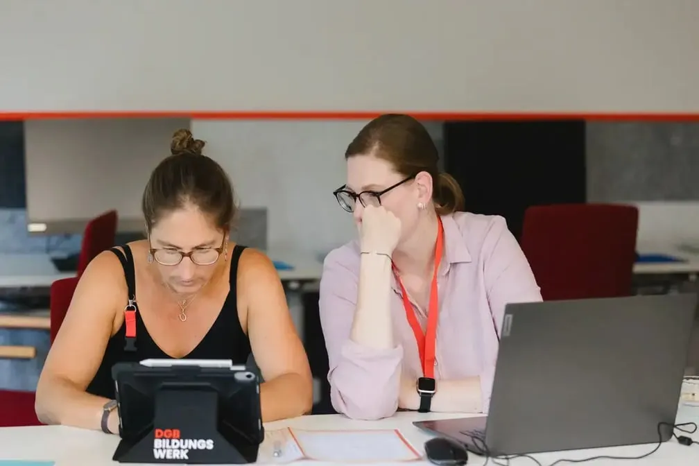 Zwei Personen arbeiten gemeinsam an einem Tisch mit Tablet und Laptop, Schriftzug „DGB Bildungswerk" auf dem Tablet sichtbar.