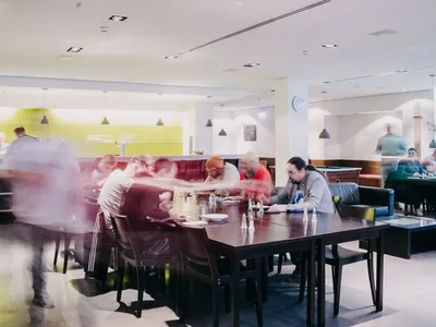 Großer Speiseraum mit langen Tischen; mehrere Personen sitzen beim Essen, einige Bewegungen sind verwischt, im Hintergrund Theke und Sitzbereiche.