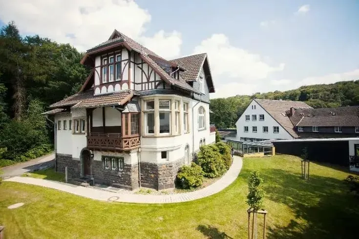 „Historisches, zweistöckiges Fachwerkhaus mit rotem Ziegeldach und Balkon, umgeben von Rasen und Bäumen; im Hintergrund weitere Gebäude unter blauem Himmel.“