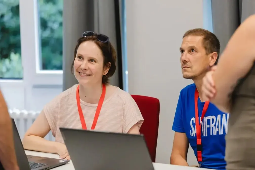 Zwei Personen sitzen an einem Tisch mit Laptops und roten Schlüsselbändern um den Hals, im Hintergrund graue Vorhänge und ein Fenster mit Blick ins Grüne.
