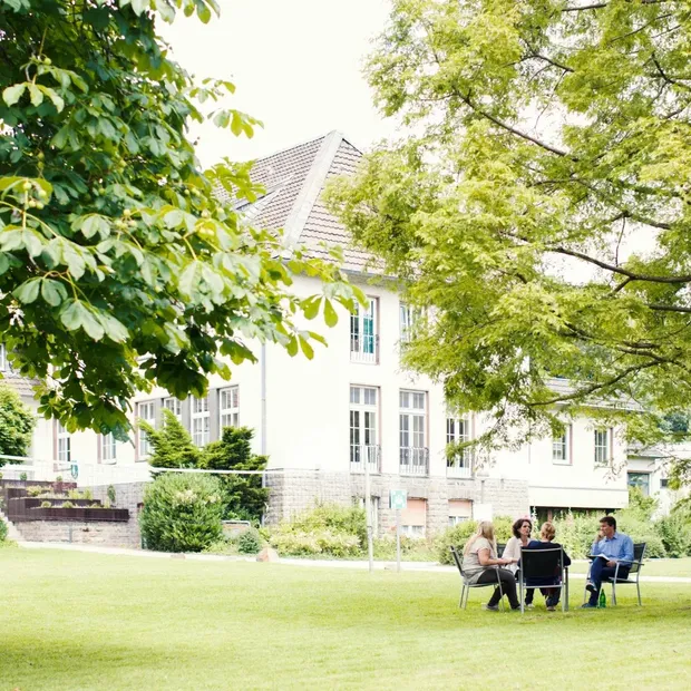Seminargruppe sitzt an einem Tisch im Grünen vor einem Bildungszentrum mit Bäumen und Rasenfläche