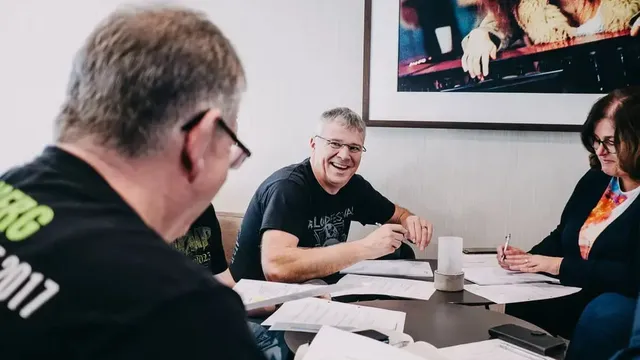 Gruppe von Personen sitzt an einem Tisch mit mehreren Dokumenten und Stiften; im Hintergrund hängt ein großes gerahmtes Bild.