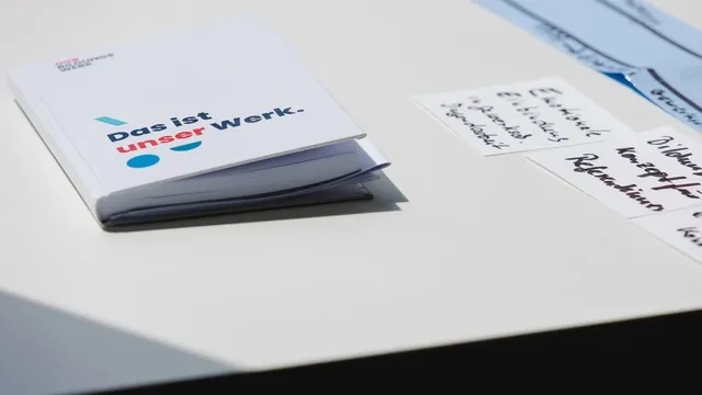 Weißes Heft mit der Aufschrift „Das ist unser Werk." liegt auf einem Tisch neben handgeschriebenen Notizzetteln.