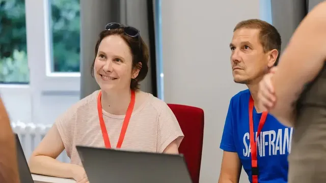 Zwei Personen sitzen an einem Tisch mit Laptops und roten Schlüsselbändern um den Hals, im Hintergrund graue Vorhänge und ein Fenster mit Blick ins Grüne.