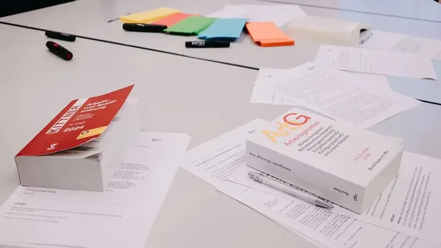 Tisch mit Fachbüchern zum Arbeitsrecht, lose Blätter, Stifte und farbige Moderationskarten in Grün, Blau, Orange und Gelb.