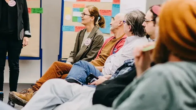 Mehrere Personen sitzen in einer Halbkreis-Runde in einem Seminarraum und hören einer stehenden Person zu. Im Hintergrund hängen Pinnwände voller bunter Moderationskarten.