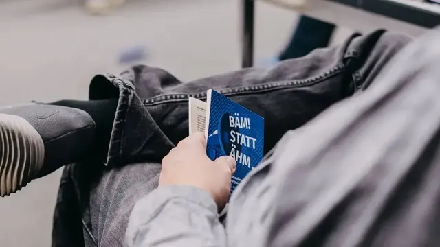 Person hält ein kleines blaues Buch mit der Aufschrift „BÄM! STATT ÄHM" in der Hand, sitzt mit überkreuzten Beinen neben einem Tisch.