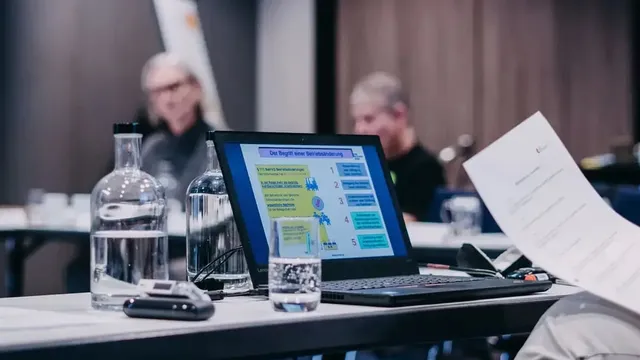 Laptop auf einem Konferenztisch zeigt eine Präsentationsfolie mit dem Titel „Der Begriff der Mitbestimmung", daneben Wasserflaschen und ein Glas.