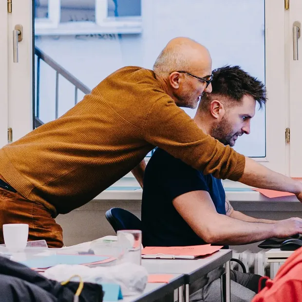 Zwei Teilnehmende arbeiten gemeinsam am Computer, eine Person erklärt etwas und zeigt auf den Bildschirm