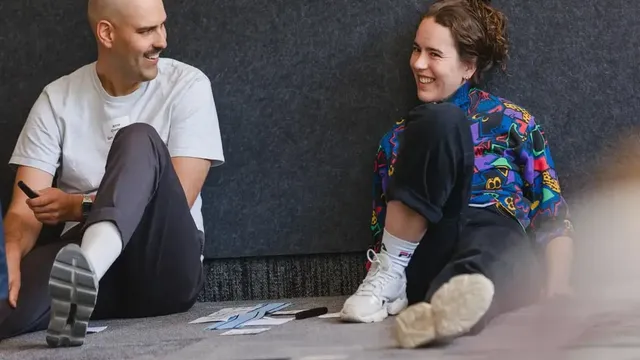 Zwei Personen sitzen auf dem Boden vor einer grauen Wand, umgeben von Papierstreifen und Stiften.