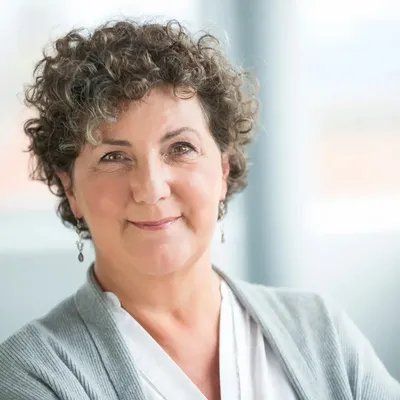 Freundlich blickende Frau (Anja Piel) in heller Kleidung vor unscharfem Hintergrund.