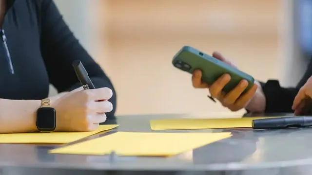 Zwei Personen sitzen an einem Tisch mit gelben Moderationskarten; eine Person hält einen schwarzen Marker, die andere ein Smartphone. Auf dem Tisch liegt zusätzlich ein weiterer Stift.
