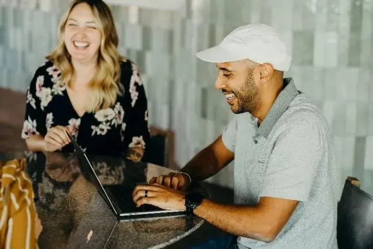 Zwei Personen sitzen an einem Tisch, eine Person arbeitet an einem Laptop, die andere sitzt daneben. Auf dem Tisch liegt eine Tasche.
