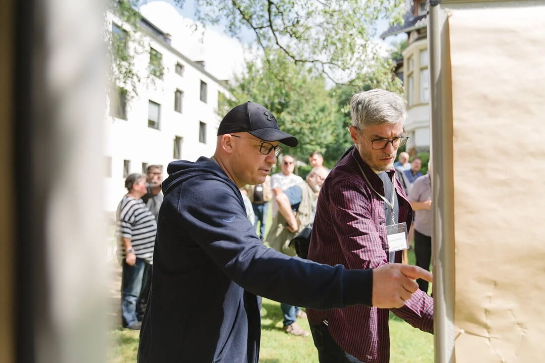 Zwei Personen stehen im Freien vor einer großen braunen Pinnwand und diskutieren, im Hintergrund weitere Menschen und Gebäude mit weißen Fassaden.