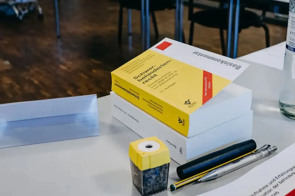 Auf einem Tisch liegen ein dicker gelber Gesetzeskommentar, ein weißes Buch, ein Bleistift, zwei Stifte und ein gelber Spitzer. Im Hintergrund stehen Tische und Stühle eines Seminarraums.