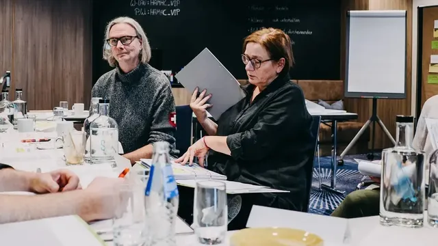 Seminarteilnehmende sitzen an einem Tisch mit Wasserflaschen, Gläsern und Unterlagen; eine Person hält einen grauen Ordner, im Hintergrund Flipchart und schwarze Wand mit Kreideaufschrift.
