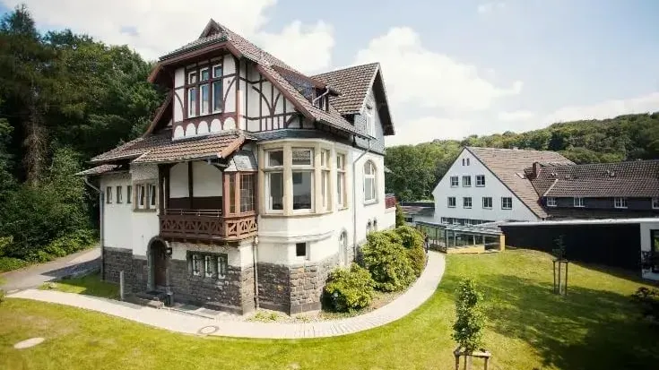 Historisches, zweistöckiges Fachwerkhaus mit rotem Ziegeldach und Balkon, umgeben von Rasen und Bäumen; im Hintergrund weitere Gebäude unter blauem Himmel.