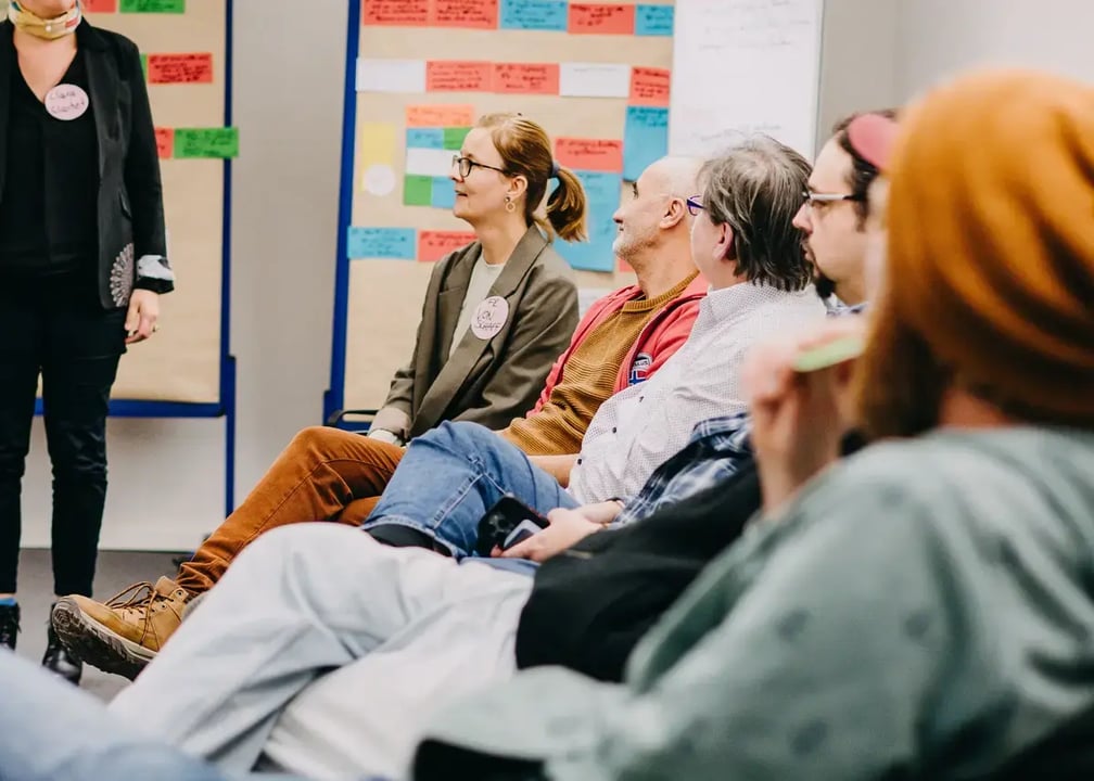 Mehrere Personen sitzen in einer Halbkreis-Runde in einem Seminarraum und hören einer stehenden Person zu. Im Hintergrund hängen Pinnwände voller bunter Moderationskarten.