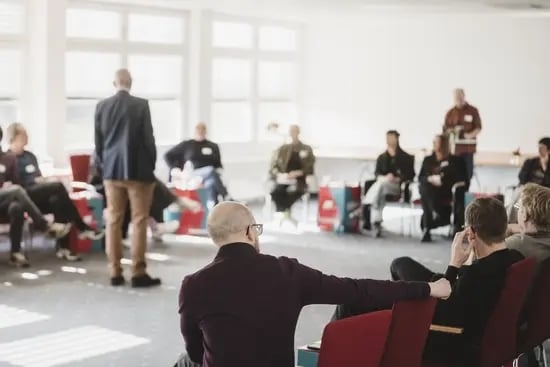 Eine größere Gruppe sitzt in einem hellen Seminarraum in einem Stuhlkreis; im Vordergrund ist eine Person auf einem roten Stuhl zu sehen, während im Hintergrund weitere Teilnehmende sowie bunte DGB‑Papphocker mit ‚#Tarifwende‘ zu erkennen sind.