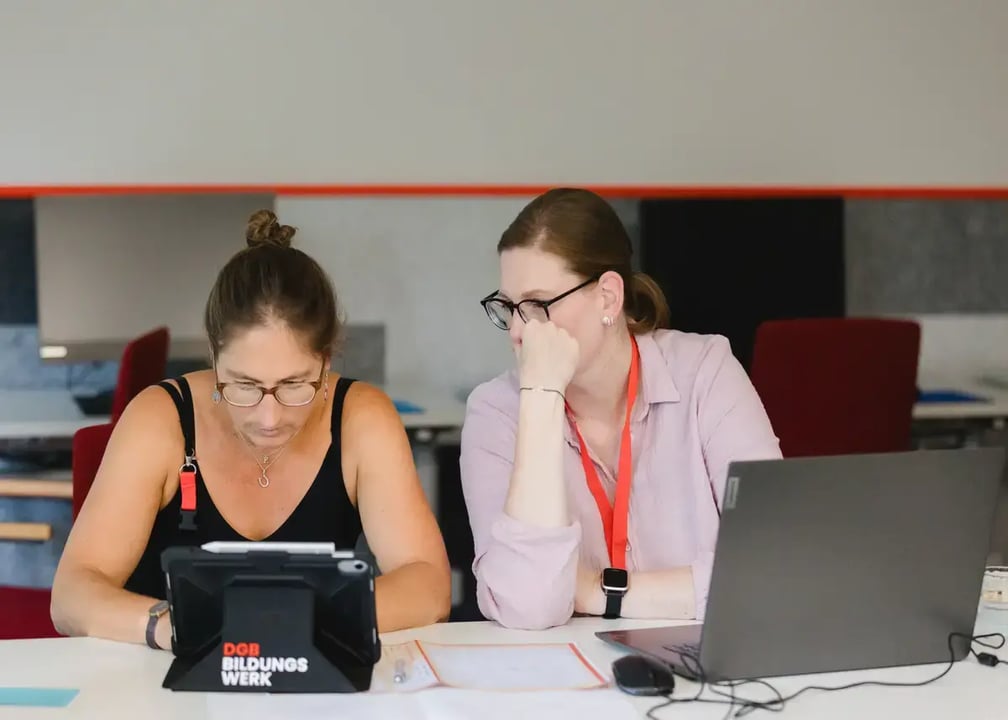 Zwei Personen arbeiten gemeinsam an einem Tisch mit Tablet und Laptop, Schriftzug „DGB Bildungswerk“ auf dem Tablet sichtbar.
