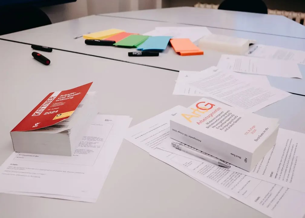 Tisch mit Fachbüchern zum Arbeitsrecht, lose Blätter, Stifte und farbige Moderationskarten in Grün, Blau, Orange und Gelb.