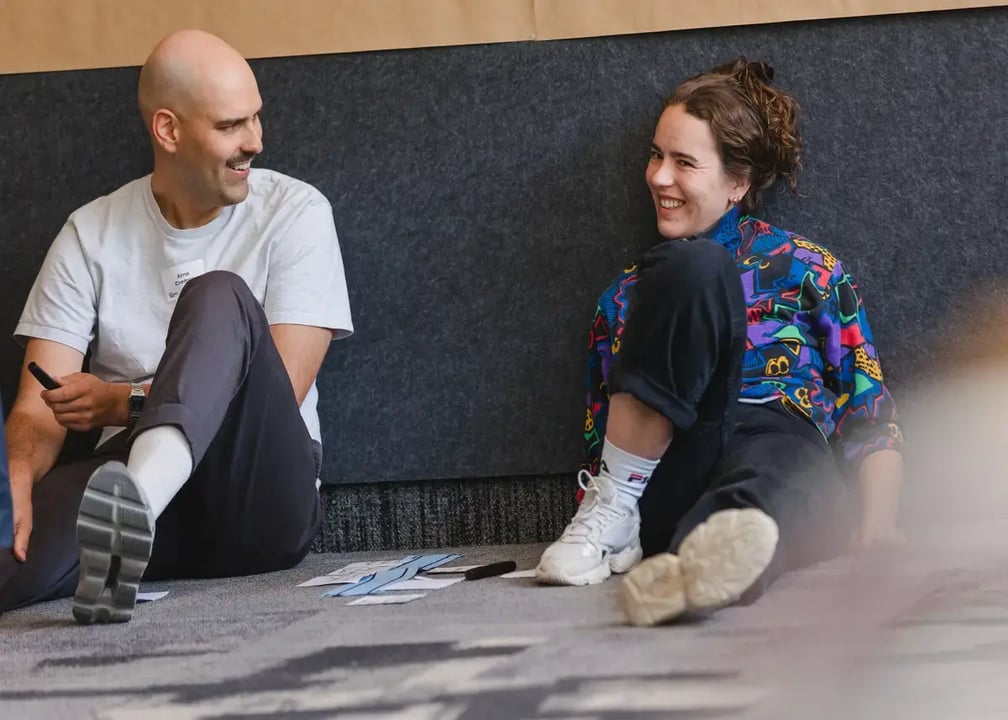 Zwei Personen sitzen auf dem Boden vor einer grauen Wand, umgeben von Papierstreifen und Stiften.