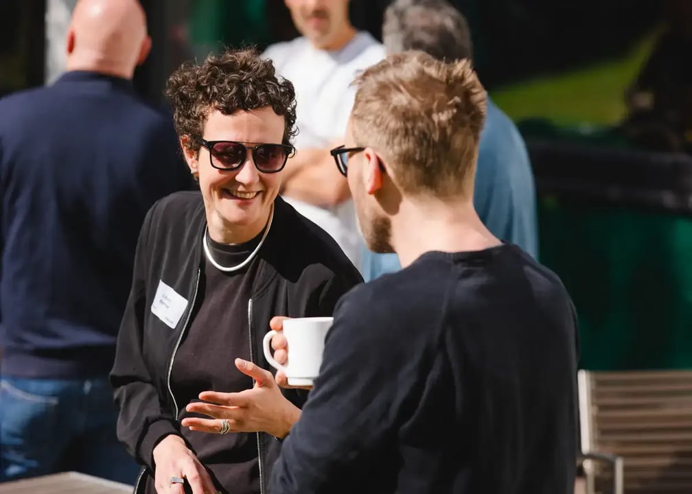 Zwei Personen im Gespräch im Freien, eine hält eine weiße Kaffeetasse, im Hintergrund weitere Personen.