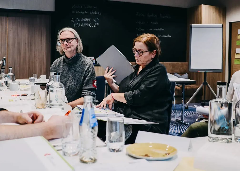 Seminarteilnehmende sitzen an einem Tisch mit Wasserflaschen, Gläsern und Unterlagen; eine Person hält einen grauen Ordner, im Hintergrund Flipchart und schwarze Wand mit Kreideaufschrift.