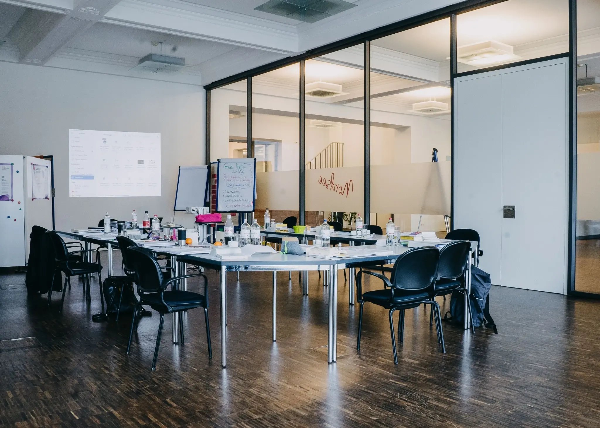 „Seminarraum mit zusammengestellten Tischen und schwarzen Stühlen; auf den Tischen stehen Getränke, Notizen und Arbeitsmaterialien, im Hintergrund Glaswände und Flipchart.“