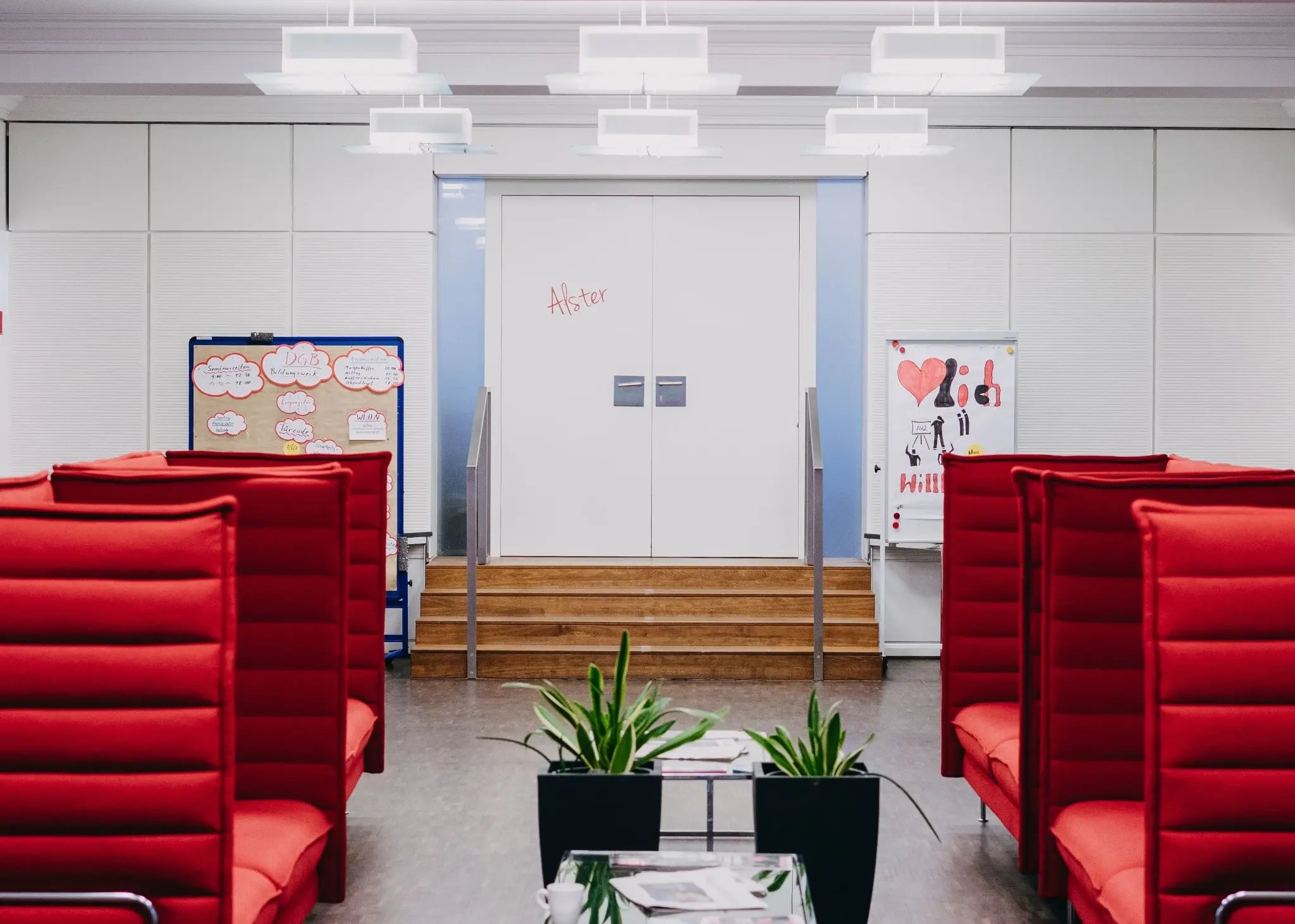 „Moderner Innenbereich mit roten Lounge-Sesseln; in der Mitte führen Stufen zu weißen Türen, daneben Pinnwände und Flipchart, vorne Pflanzen auf einem Tisch.“