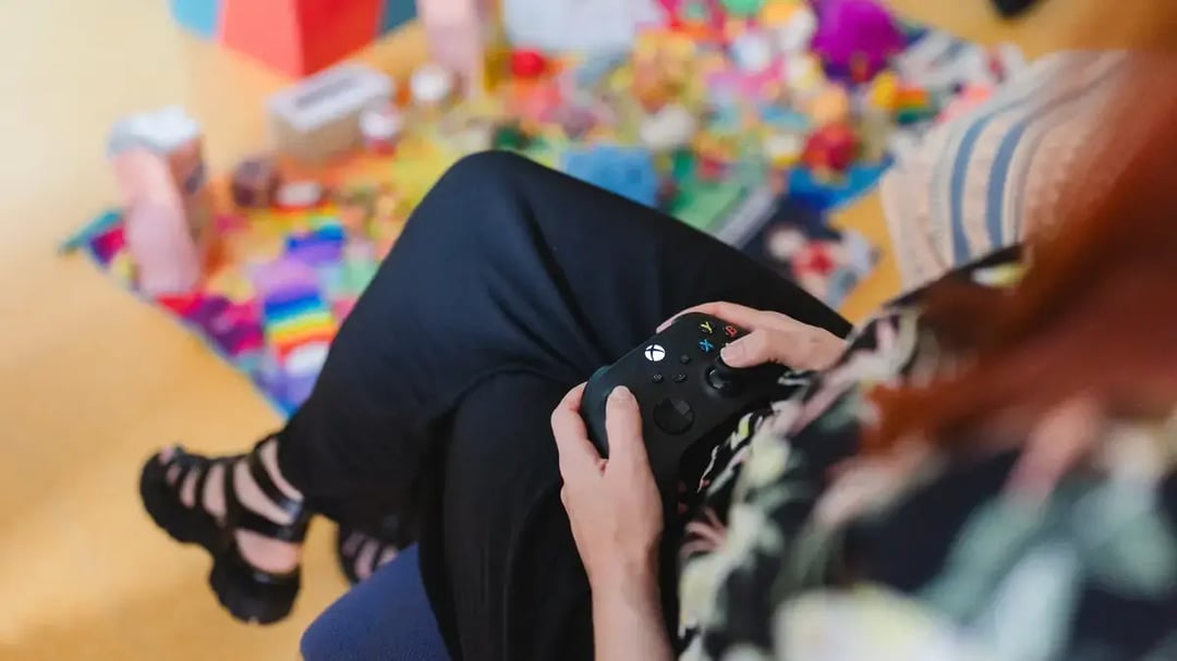 Eine Frau hält einen Gamecontroller in den Händen und spielt vor einem leicht verschwommenen, bunt beleuchteten Hintergrund.