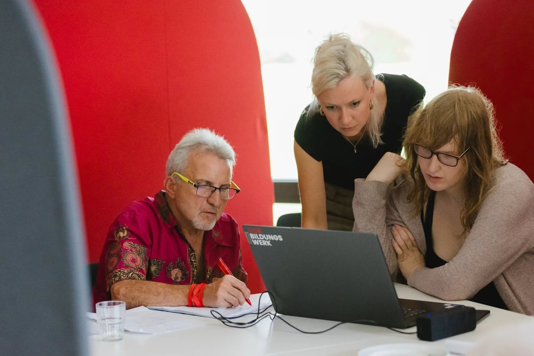 Drei Personen arbeiten gemeinsam an einem Laptop mit der Aufschrift „Bildungswerk“, daneben liegen Unterlagen und ein Glas Wasser; rote Raumtrenner im Hintergrund.