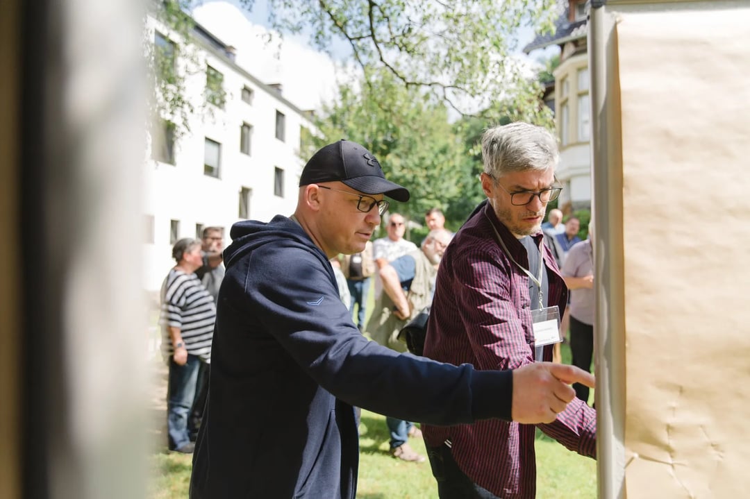 Zwei Personen stehen im Freien vor einer großen braunen Pinnwand und diskutieren, im Hintergrund weitere Menschen und Gebäude mit weißen Fassaden.