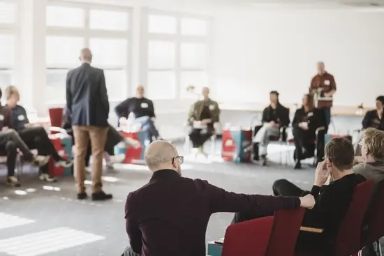 Eine größere Gruppe sitzt in einem hellen Seminarraum in einem Stuhlkreis; im Vordergrund ist eine Person auf einem roten Stuhl zu sehen, während im Hintergrund weitere Teilnehmende sowie bunte DGB‑Papphocker mit ‚#Tarifwende‘ zu erkennen sind.