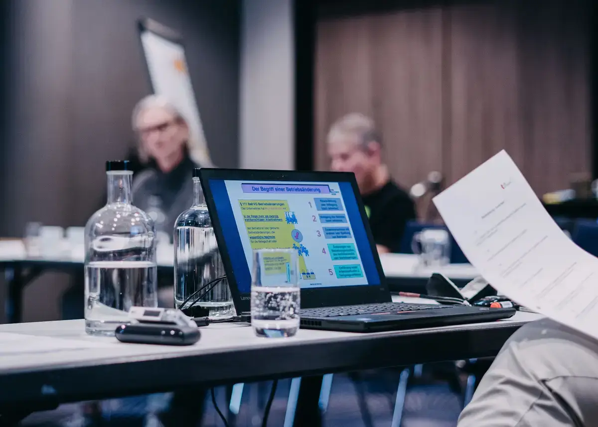 Laptop auf einem Konferenztisch zeigt eine Präsentationsfolie mit dem Titel „Der Begriff der Mitbestimmung“, daneben Wasserflaschen und ein Glas.