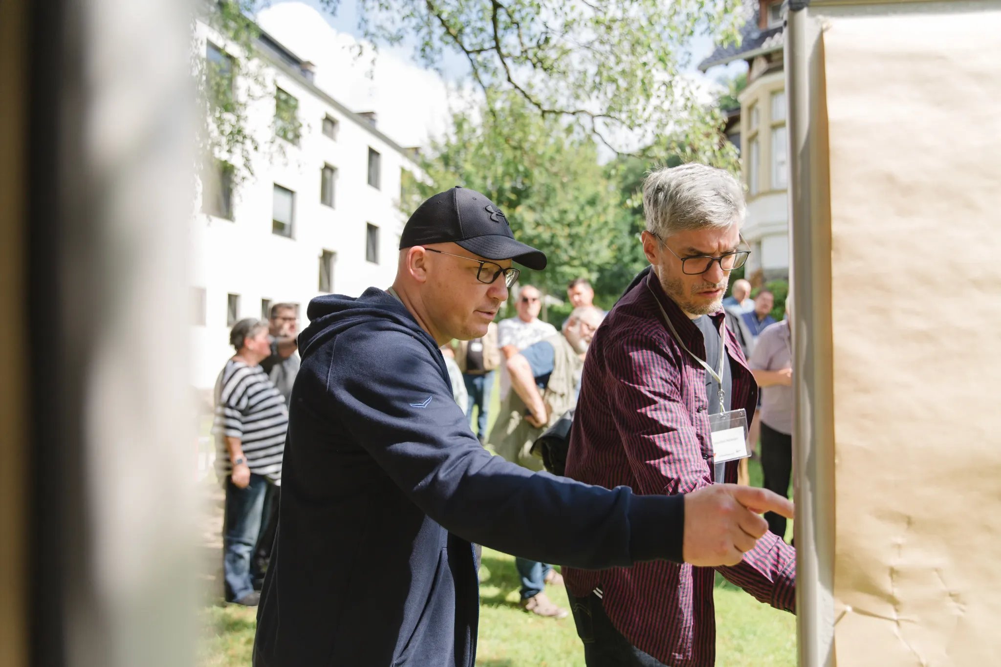 Zwei Personen stehen im Freien vor einer großen braunen Pinnwand und diskutieren, im Hintergrund weitere Menschen und Gebäude mit weißen Fassaden.