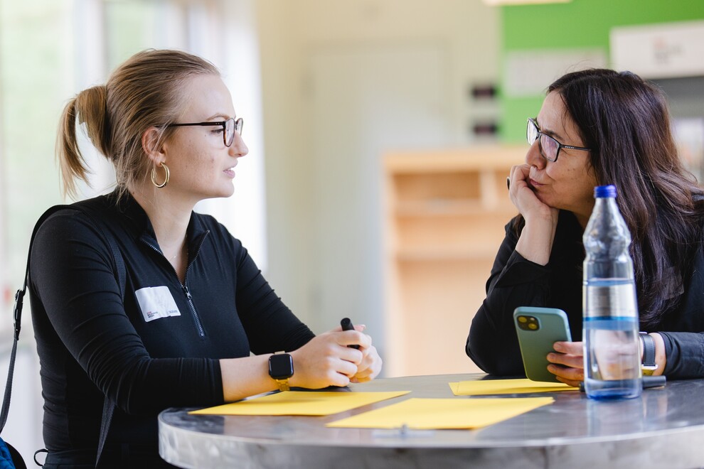 Zwei Frauen sitzen an einem runden Tisch und führen ein Gespräch. Auf dem Tisch liegen gelbe Karten, eine Frau hält ein Handy, die andere einen Stift.
