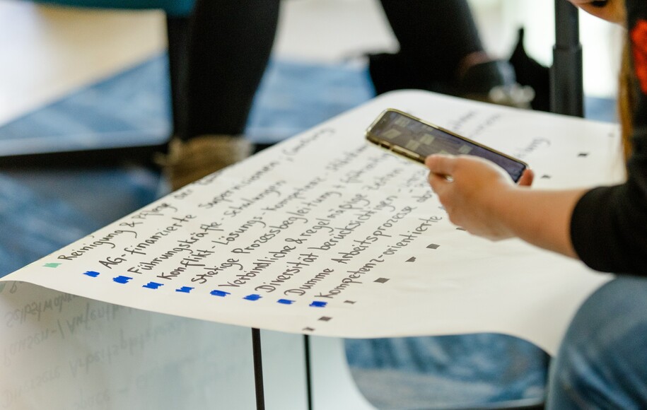 Eine Person hält ein Smartphone in der Hand und blickt auf ein Blatt Papier oder eine Tafel mit handgeschriebenem Text in Listenform.