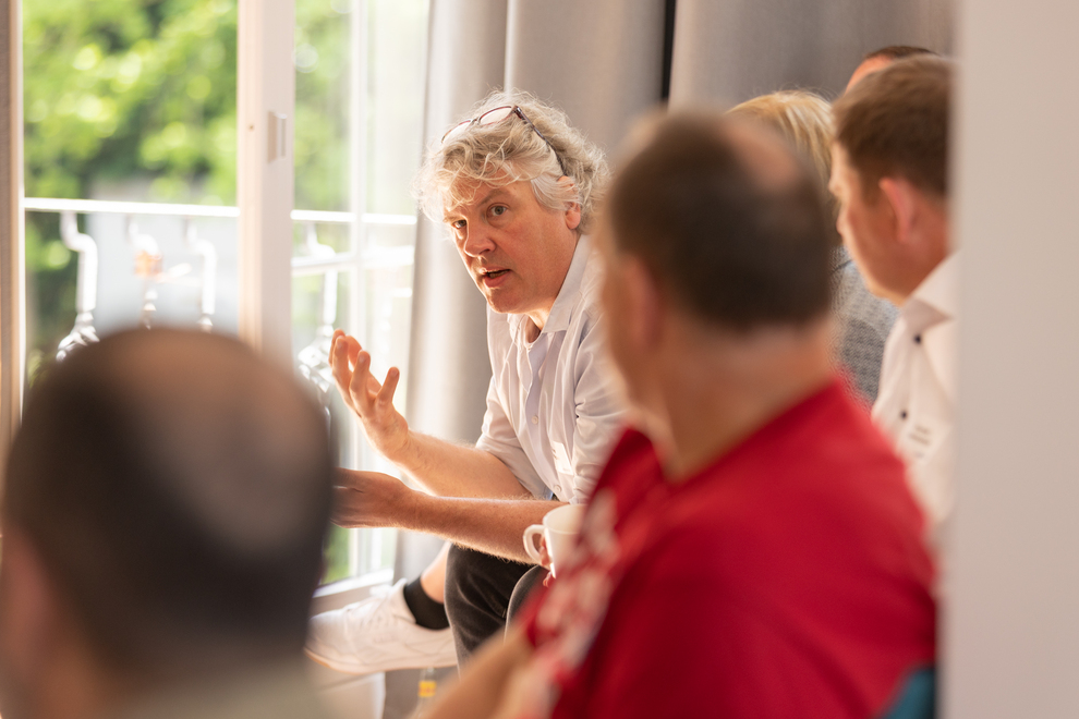 Eine Gruppe von Personen sitzt in einem Raum und führt eine Diskussion. Eine Person in der Mitte gestikuliert beim Sprechen.