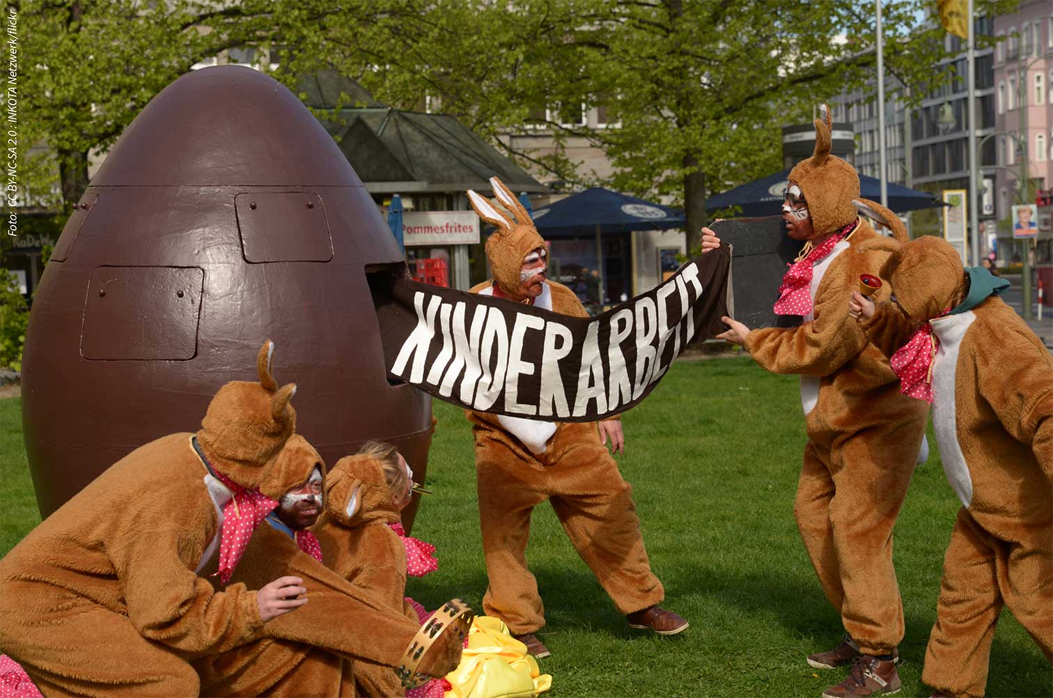 Eine Gruppe von Menschen, verkleidet als Osterhasen, vor einem riesigen schokoladenbraunen Ei in Form einer Riesen-Handgranate. Einer der Verkleideten zieht aus dem Ei ein Transparent auf dem steht: KINDERARBEIT