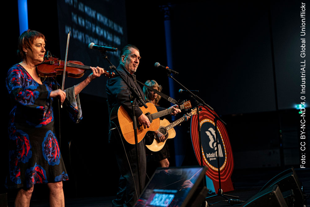 Ein musikalisches Trio mit 2 Gitarren und 1 Violine, rechts daneben im Bild: Das Logo von IndustriALL Global Union