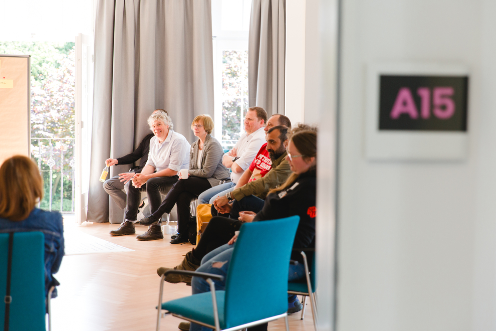 Eine Gruppe von Personen sitzt in einem Raum mit großen Fenstern und grauen Vorhängen. An der Wand hängt ein Schild mit der Aufschrift „A15“ in pinker Schrift.