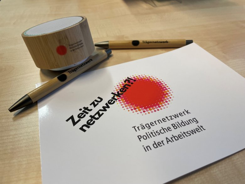 Eine Postkarte, zwei Stifte und eine Lautsprecherbox mit dem Logo des Projektes Trägernetzwerk liegen auf einem Tisch.